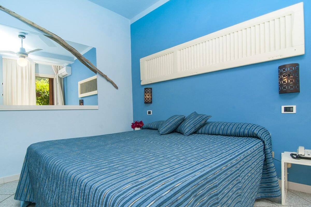 Une chambre à coucher avec un lit, une fenêtre, un ventilateur de plafond, un radiateur et une petite table sur laquelle se trouve un téléphone.