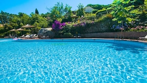 Une piscine d’un bleu éclatant, bordée de chaises longues et d’ombrelles, entourée d’une végétation luxuriante et d’un mur de pierre.