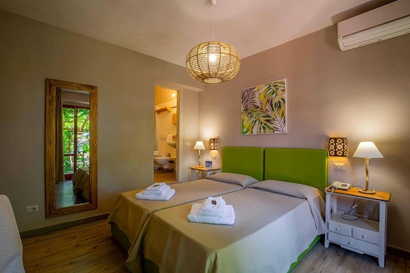 A hotel room with two beds, a mirror, a green headboard, a wall art piece, and a bedside table with a lamp.
