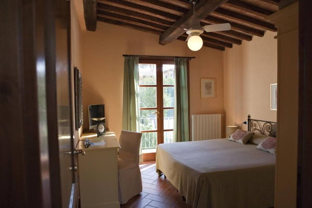 Une chambre à coucher comprenant un lit, une chaise, un bureau avec une horloge, une fenêtre avec des rideaux et un ventilateur de plafond.