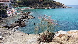 A coastal area with rocky terrain, clear blue water, and a beach with umbrellas.