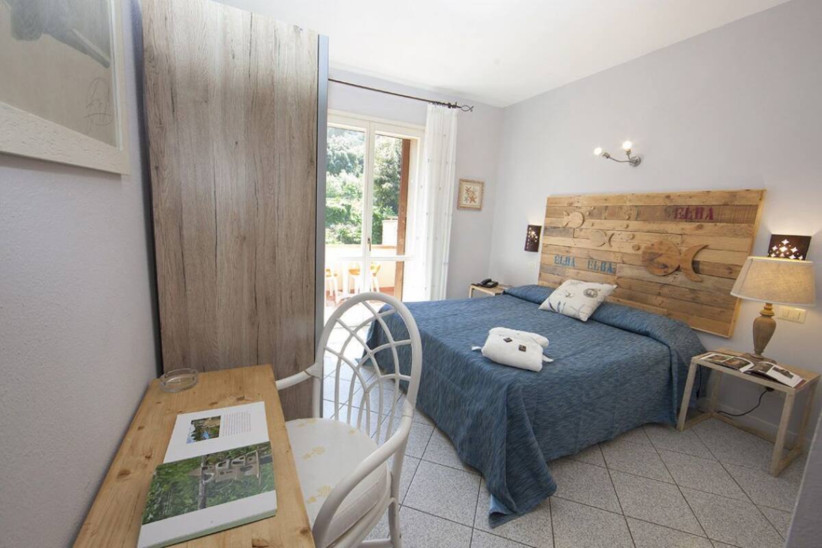 Une chambre à coucher avec une tête de lit en bois, un lit avec une literie bleue, une chaise blanche, une petite table avec un livre et une table de chevet avec une lampe.