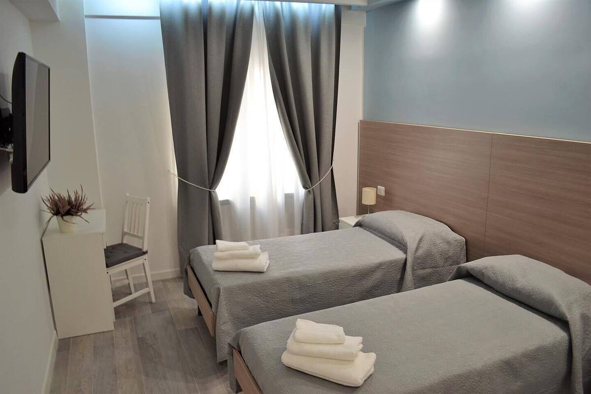 Ein Hotelzimmer mit zwei Betten, einem Sessel, einem Fernseher und einem Fenster mit Vorhängen.