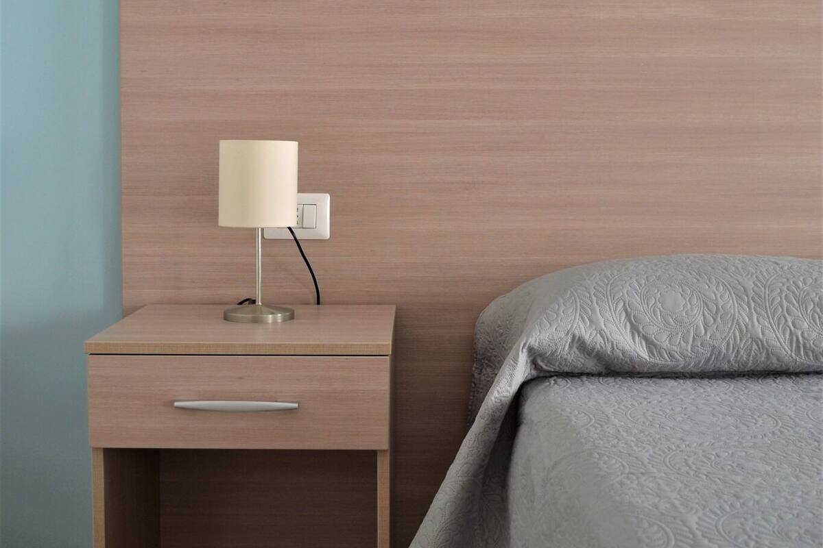 Ein ordentlich bezogenes Bett mit grauer Decke, ein Nachttisch mit Lampe und eine Schublade.