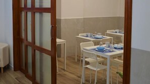 Una habitación con puerta corrediza de vidrio que da a un comedor con mesas y sillas blancas, un taburete blanco pequeño y una planta en maceta.