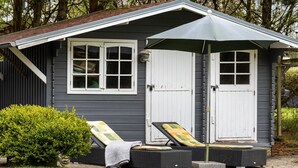 Eine kleine, dunkelgraue Gartenhütte mit weißen Verzierungen, ein überdachter Patio-Bereich mit zwei Liegestühlen und einem Tisch sowie ein ordentlich geschnittener Busch davor.