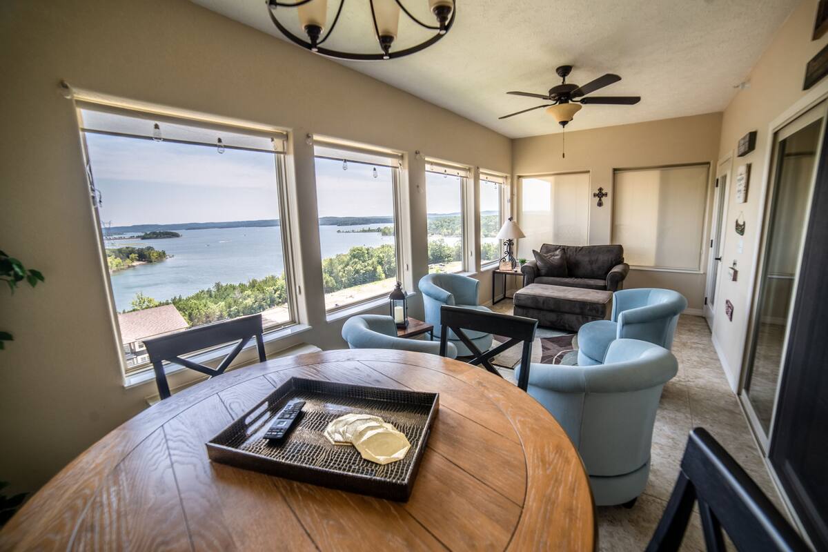 Quarto espaçoso com uma grande janela que oferece vista para um lago e terras distantes. Há uma mesa de madeira redonda com um tabuleiro e controles remotos. Poltronas azul-claras e um sofá escuro estão dispostos em torno de uma mesa de centro.