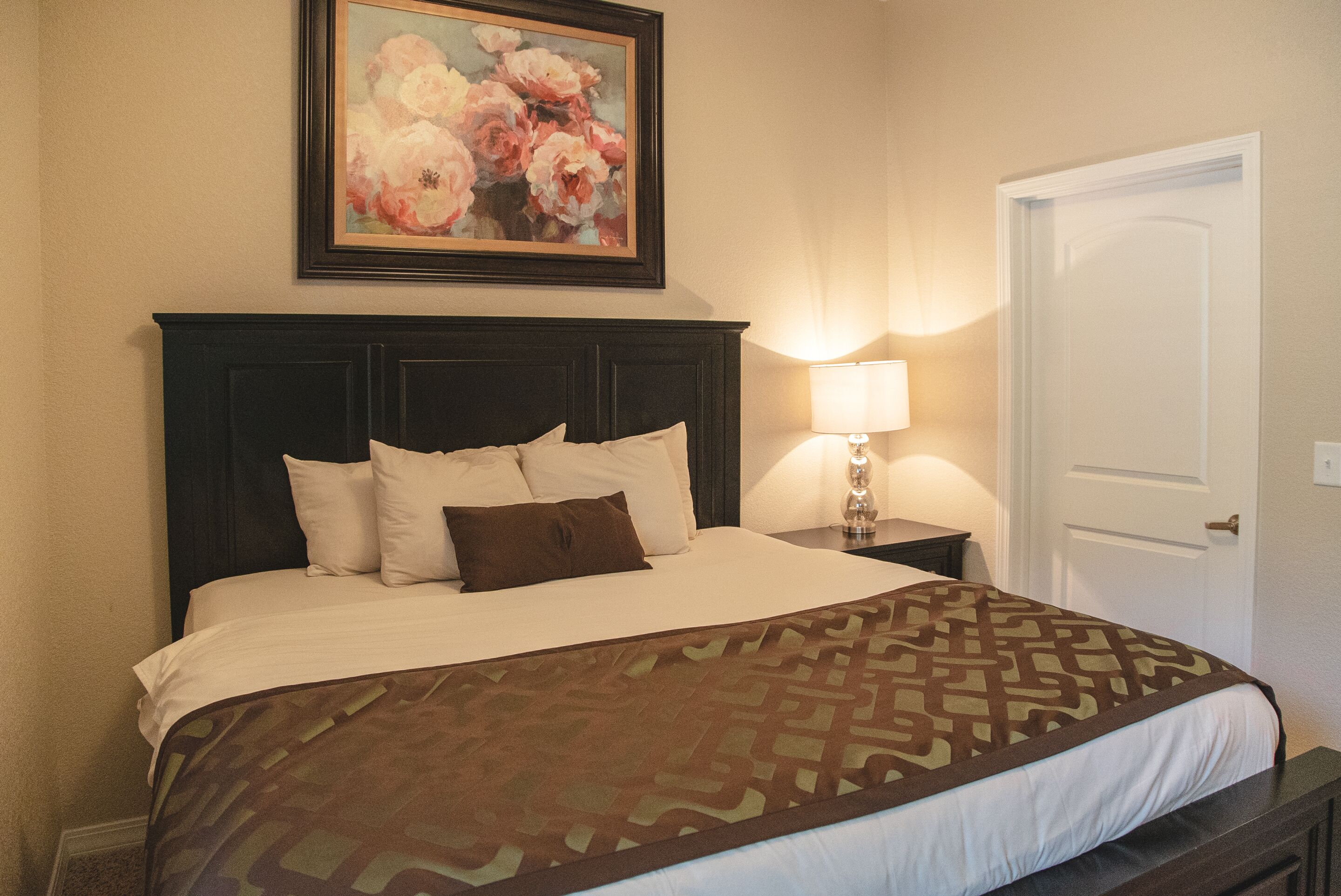 Un lit bien fait, avec une couvre-lit à motifs, une tête de lit sombre et un tableau floral encadré au-dessus.