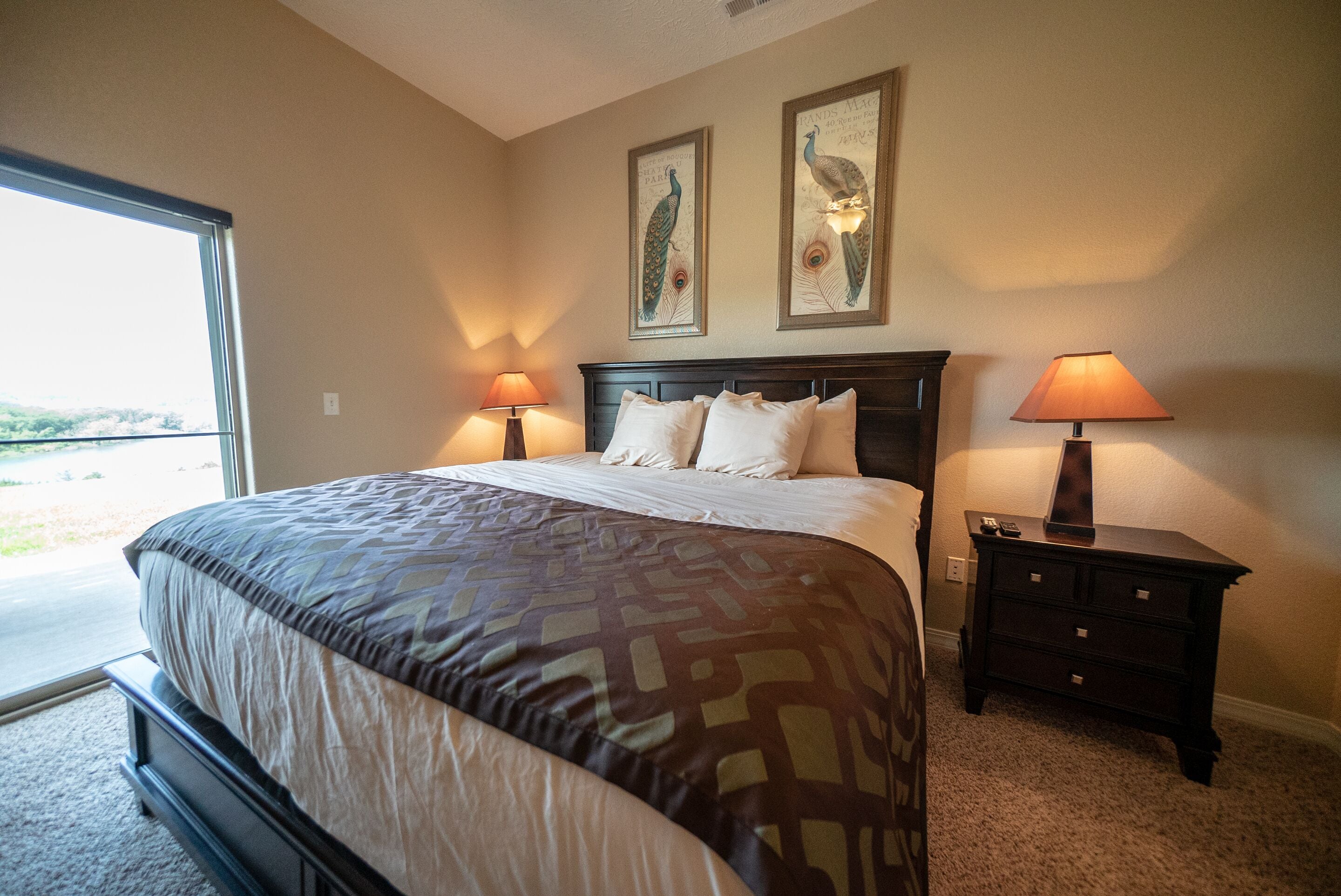 A bedroom with a large bed, two wall-mounted lamps, a nightstand, and two framed peacock pictures.