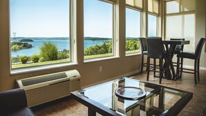 Ein Zimmer mit großen Fenstern, das einen Blick auf einen See und grüne Vegetation bietet. Es gibt einen Tisch mit Stühlen und einen Glastisch.