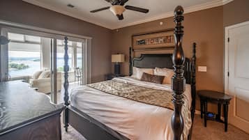 A bedroom with a four-poster bed, a ceiling fan, a nightstand with a lamp, and a view of a body of water through large windows.