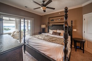 A bedroom with a four-poster bed, a ceiling fan, a nightstand with a lamp, and a view of a body of water through large windows.