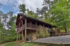 A rustic cabin with a wooden deck and stone foundation, surrounded by trees.