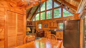 Interior de una cabaña de madera con una mesa de comedor, sillas, un refrigerador y una chimenea de piedra.
