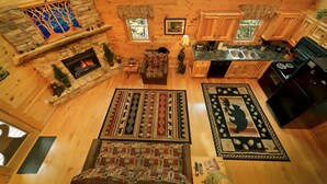 Interior de una cabaña con chimenea de piedra, paredes de madera, cocina con pileta y un ventanal grande con vista al bosque.