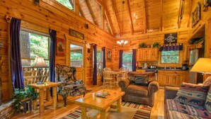 A cozy cabin interior with wooden walls, a dining area, a living room with a sofa and chairs, and a kitchen with wooden cabinets.