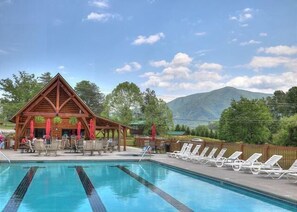Un área de piscina con sillones de descanso, un edificio de madera con techo y montañas al fondo.