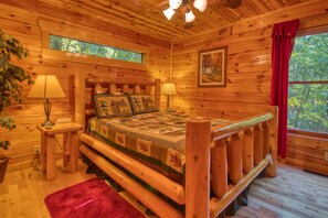 Un dormitorio de cabaña de madera con una cama, dos lámparas de noche, un ventanal con cortina roja y un cuadro en la pared.