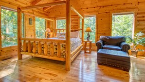 A log cabin bedroom with a four-poster bed, a leather sofa, and large windows offering a view of the forest.