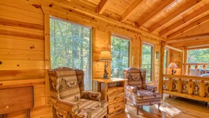 A cozy cabin interior with wooden walls and floors, two patterned armchairs, a small table with a lamp, and large windows offering a view of the surrounding trees.