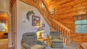 A cozy room with a wooden staircase, two armchairs, a lamp, and framed pictures on the wall.