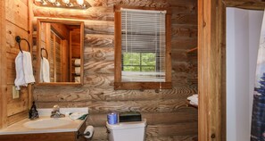 Une salle de bain rustique, aménagée dans une cabane en bois, comprenant un lavabo, un miroir, des toilettes et une fenêtre avec des stores.