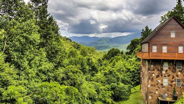 A rustic stone house with a balcony, surrounded by lush green forest and mountains in the background. - Hillbilly Hilton #525 (Gatlinburg)