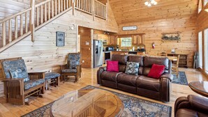 A cabin interior with a wooden staircase, a living room with a leather sofa and chairs, a kitchen area, and a dining table. - Hillbilly Hilton #525 (Gatlinburg)