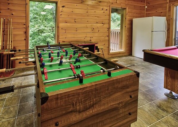 A foosball table in a wooden room with a pool table and a refrigerator.