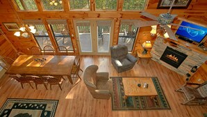 Geräumiges Blockhauskabinen-Interieur mit Essbereich, Kamin, Fernseher und großem Fenster mit Blick auf eine Terrasse.