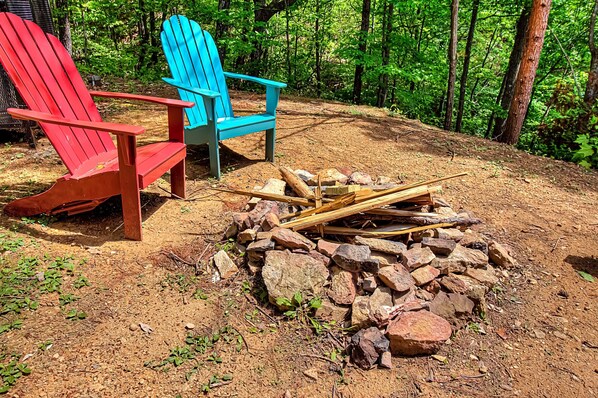 Two Adirondack chairs, one red and one blue, placed on a dirt ground with a pile of firewood and rocks nearby.