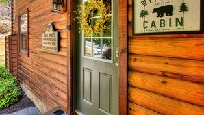 Una cabaña con fachada de madera, una puerta verde, un cartel de bienvenida con la silueta de un oso y un adorno colgando sobre la puerta.