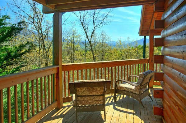 A wooden deck with two wicker chairs and a small table, overlooking a forested landscape with mountains in the distance.