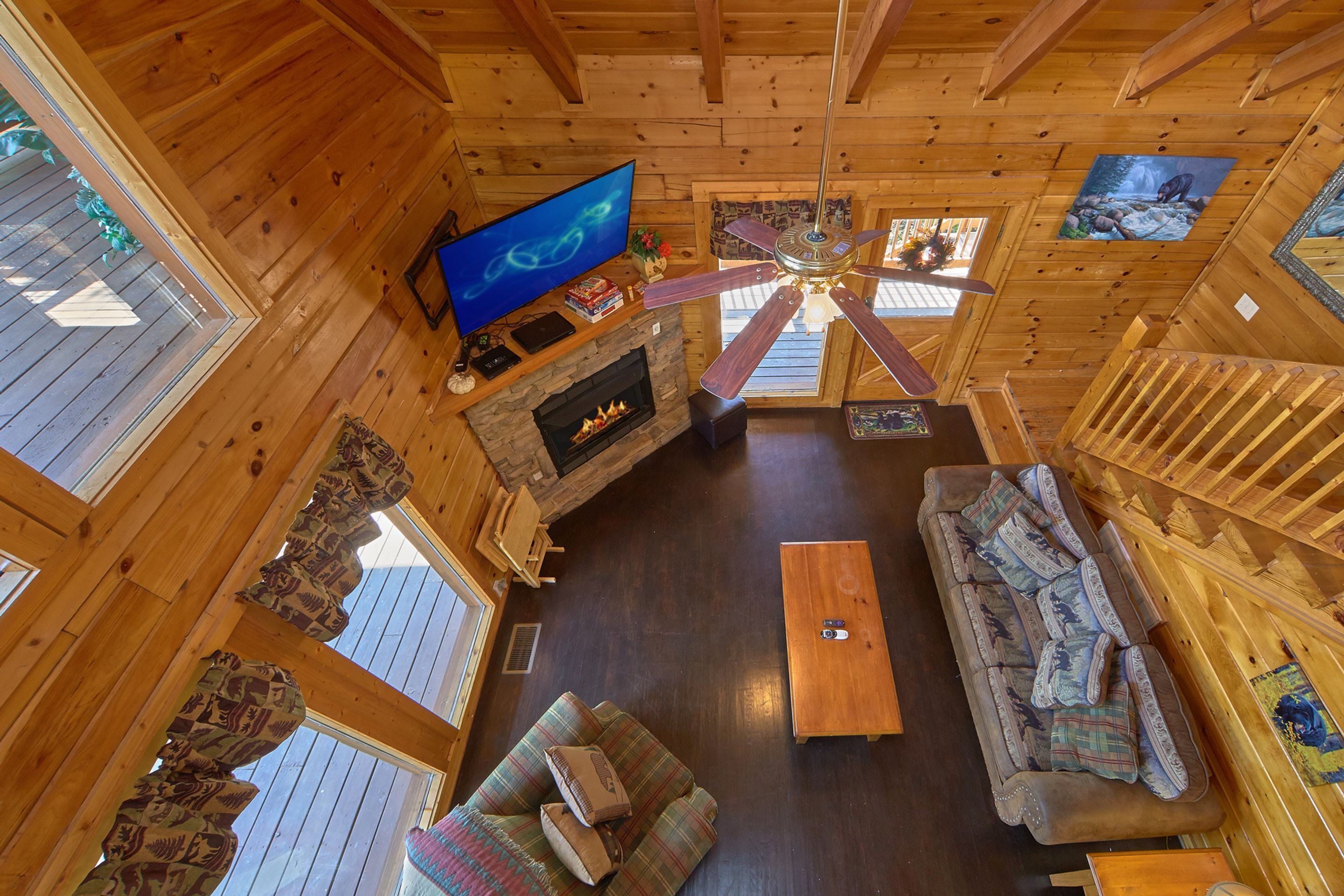 Interior de una cabaña de madera con chimenea, televisor, ventilador de techo, sofá y escalera de madera.