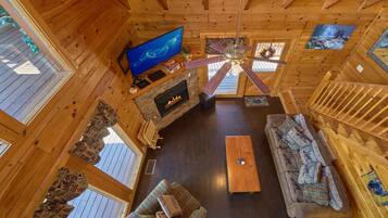 Interior de una cabaña de madera con chimenea, televisor, ventilador de techo, sofá y escalera de madera.