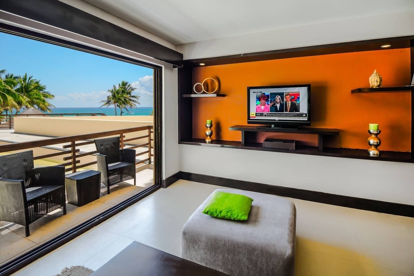 Un salon moderne doté d’une grande fenêtre donnant sur une plage, d’un téléviseur à écran plat et d’un coin salon confortable agrémenté d’un coussin vert.