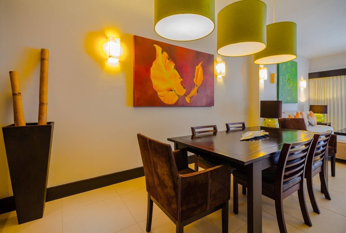 A dining area with a long table, chairs, and hanging green lamps. There is a painting on the wall and a potted plant with bamboo stalks.