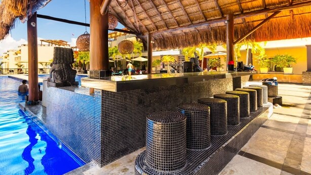 Zona de piscina con pared de azulejos, techo de paja y barra con taburetes.