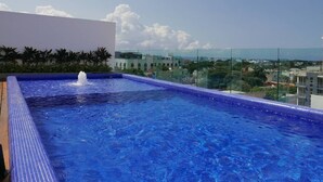 A rooftop infinity pool with a water fountain, surrounded by glass railings and overlooking a cityscape. - 3-br Condo, Partial Ocean View, 2blocks From Ocean (Playa del Carmen)
