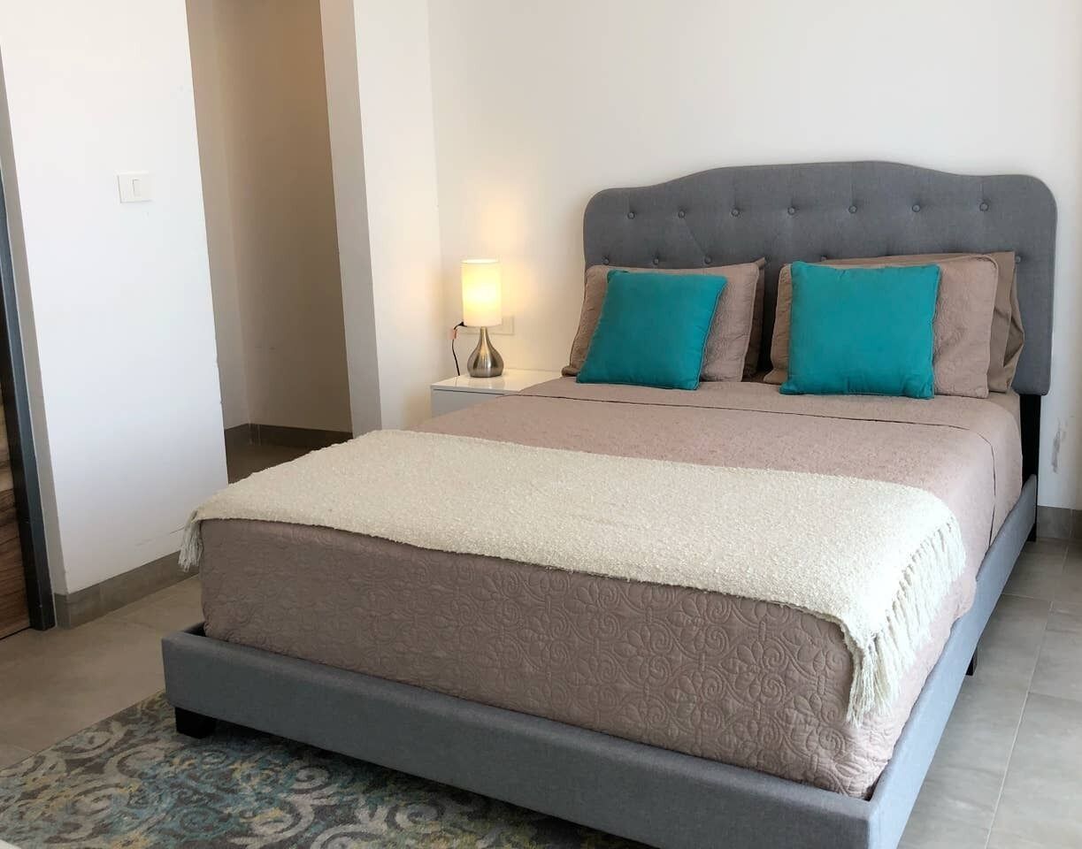 Una cama con cabecera tapizada en gris, colcha beige y blanca, y dos almohadas color turquesa.