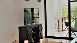 Un coin repas avec une table en bois dressée pour quatre personnes, un téléviseur posé sur un meuble et une grande fenêtre donnant sur l’extérieur.