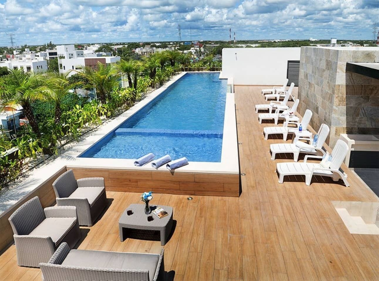 A rooftop pool area with lounge chairs and a small table, surrounded by greenery and buildings in the background.
