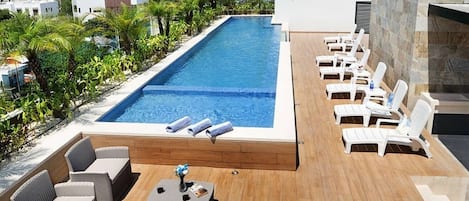 A rooftop pool area with lounge chairs and a small table, surrounded by greenery and buildings in the background.