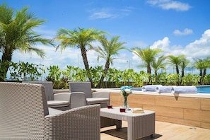 An outdoor seating area with wicker furniture, a pool, and palm trees.