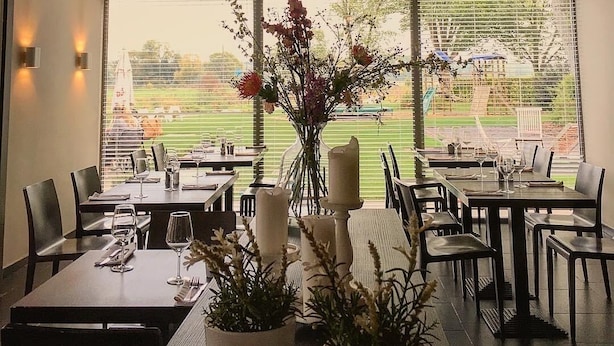 A restaurant with round, woven pendant lights, a view of a golf course through large windows, and tables set with wine glasses and candles. - Hotel La Passerelle (Durbuy)