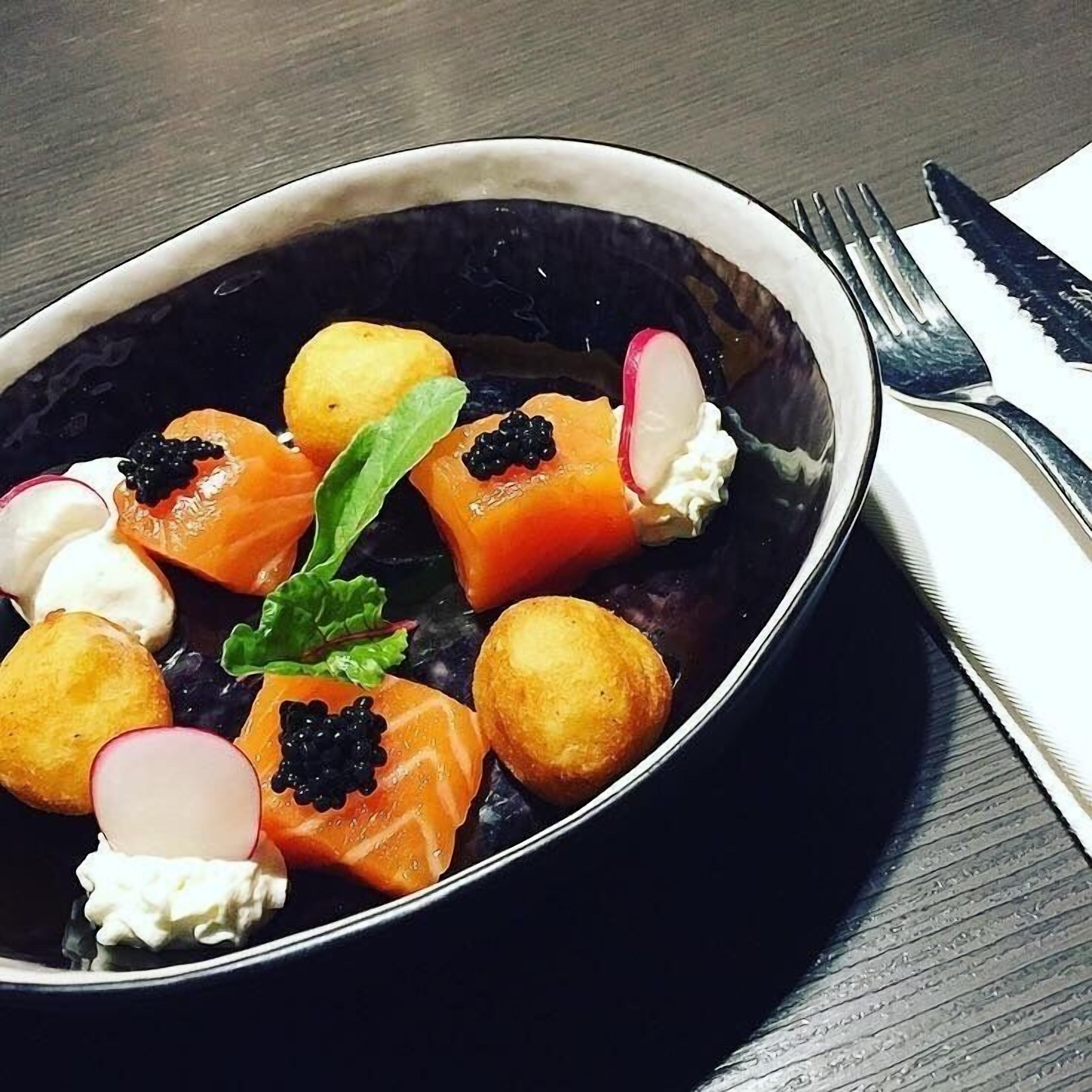 Un plato oscuro con un bol de comida que incluye salmón, caviar, rábanos y una guarnición.