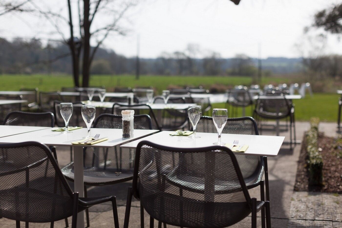 An outdoor dining area with tables and chairs, each set with a wine glass and a salt shaker.