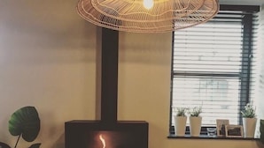 A cozy room with a wood-burning stove, a large woven umbrella light, and a window with potted plants. - Hotel La Passerelle (Durbuy)