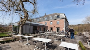 An outdoor dining area with tables and chairs, a brick building, and a glass structure. - Hotel La Passerelle (Durbuy)