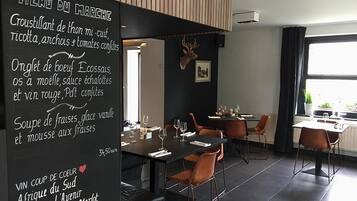 Een restaurantinterieur met een zwart menubord waarop gerechten en prijzen staan vermeld, houten lambrisering aan het plafond en een groot raam met uitzicht op groen.